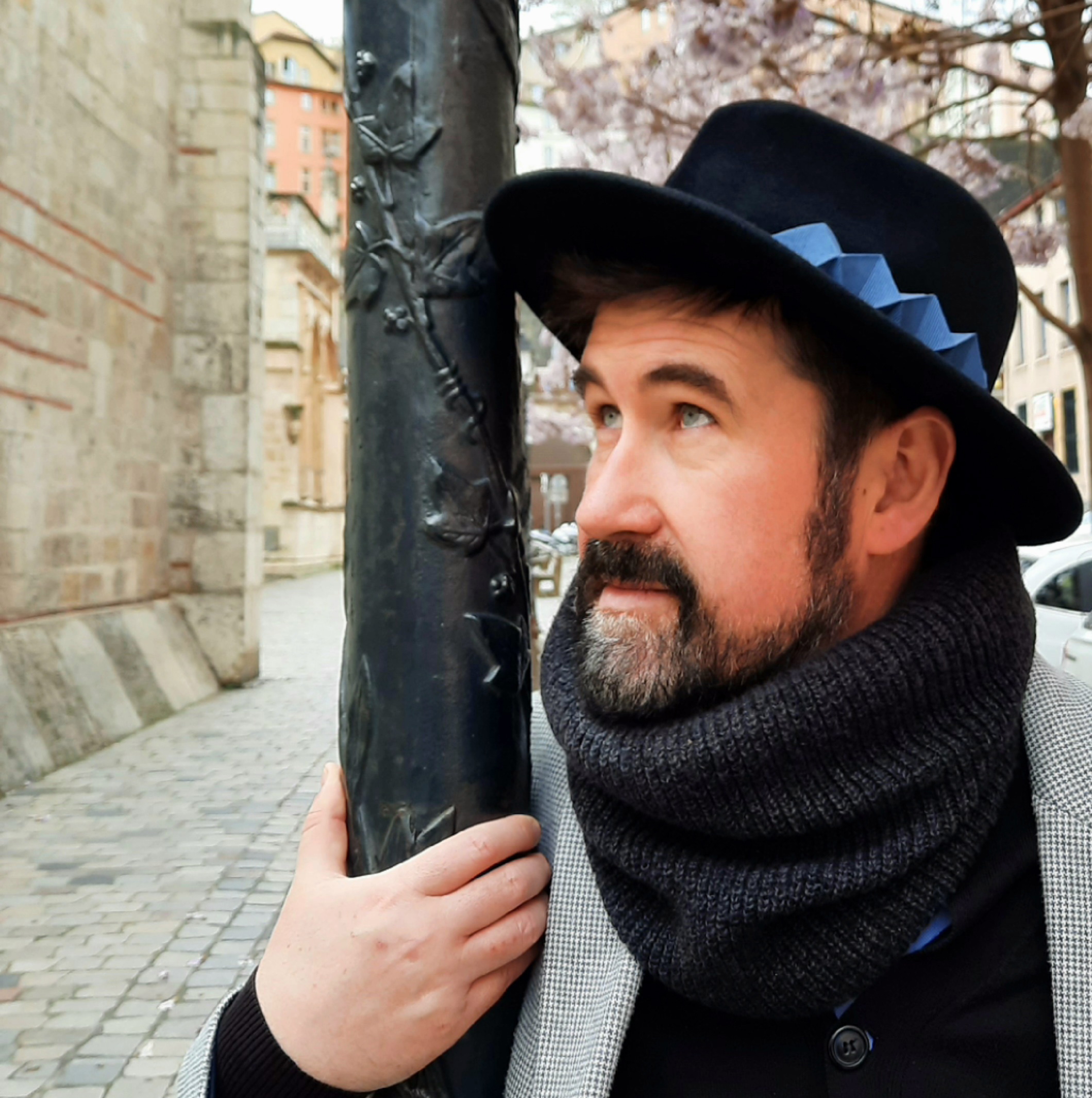 A handsome man in a black fedora with a blue origami ornament. The hat, his cardigan and his blazer create a casual city look