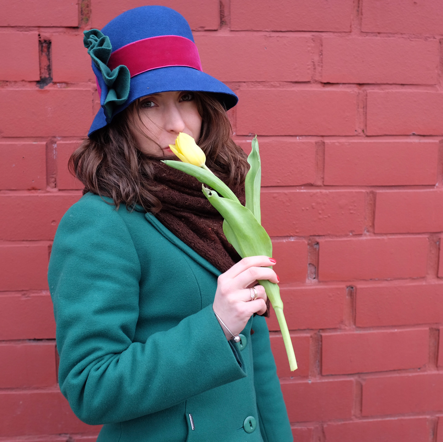 A woman with a yellow tulip wearing a custom made blue cloche hat with a big green detail matching perfectly her green coat.