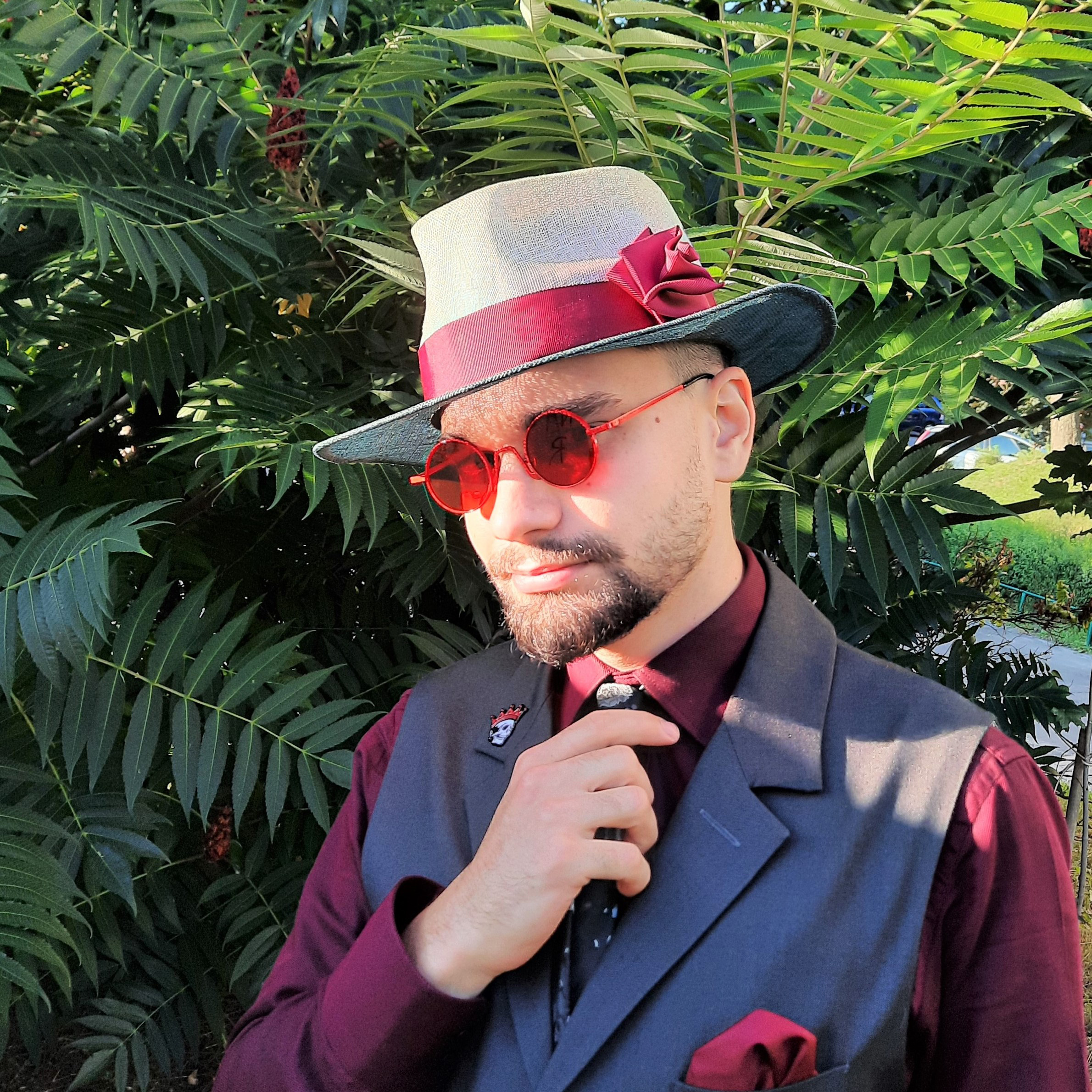 A handsome man in a white and blue summer fedora with a burgundy 3D detail which matches his waistcoat and a burgundy shirt.