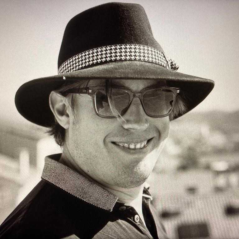 A handsome man in a bespoke fedora hat with a houndstooth ribbon. His sunglasses and a big smile complete the perfect look.