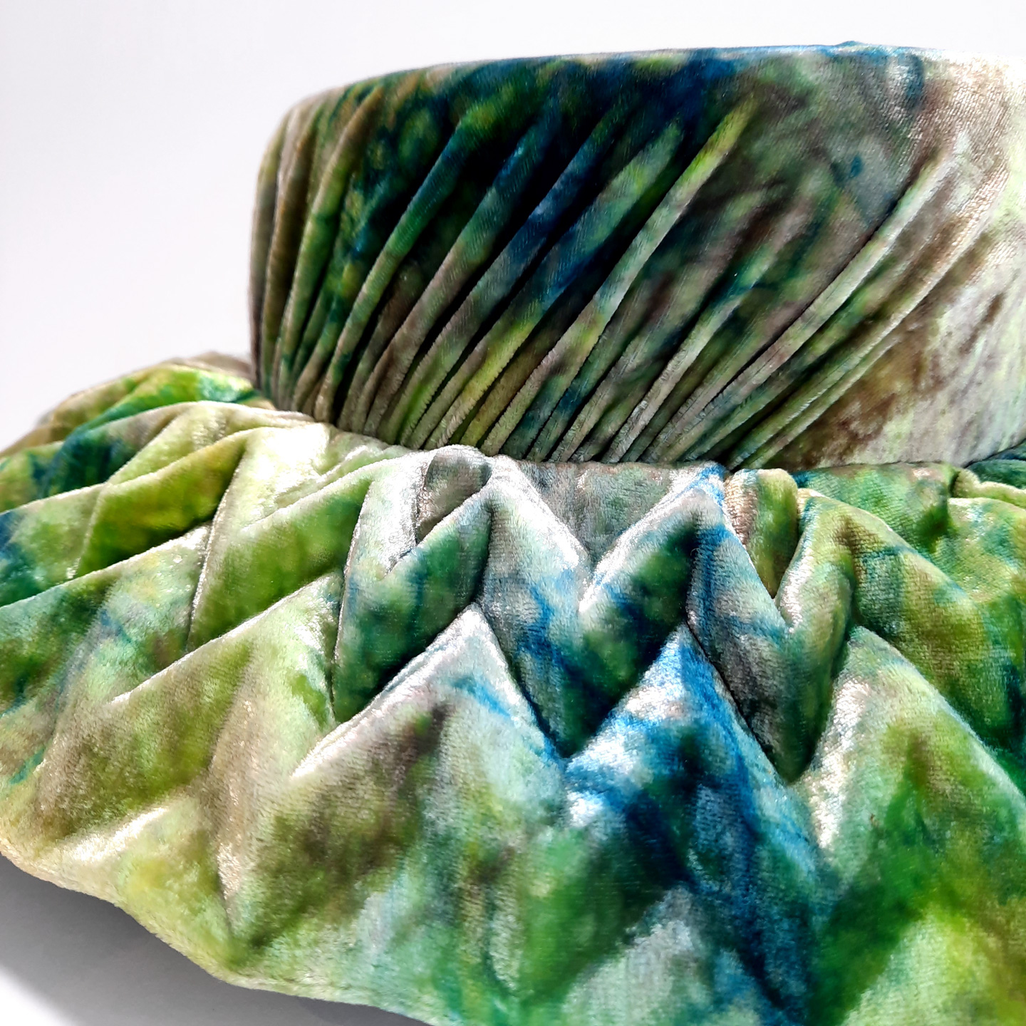 A close-up on an intricately pleated origami brim of a designer hat. The velvet silk it's made of dazzles with colors.