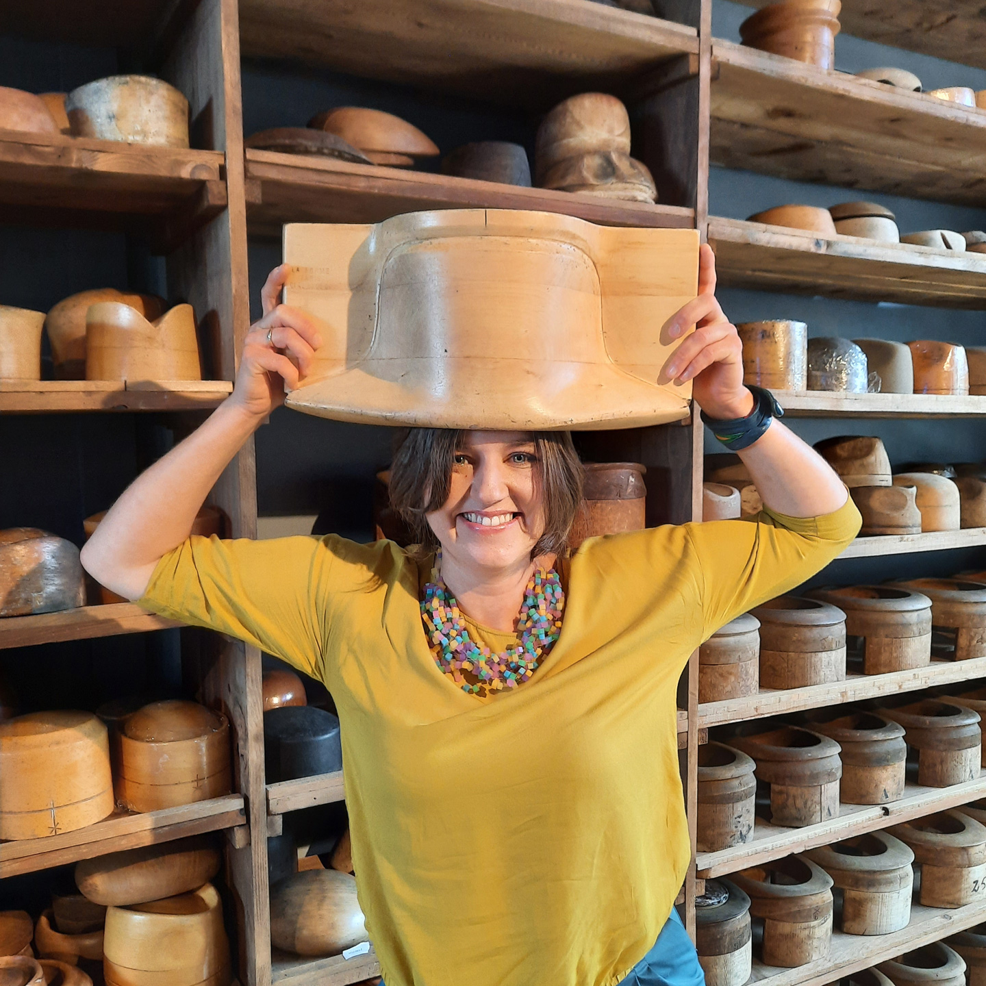 Fusanna, the hatmaker who designs and creates unique hats, holding a big hatblock over her head. Hatblocks in the background.