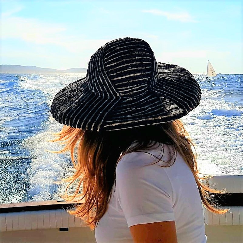 A woman on a yacht at sea in a made to measure, large brimmed summer hat and her hair floating in the wind.