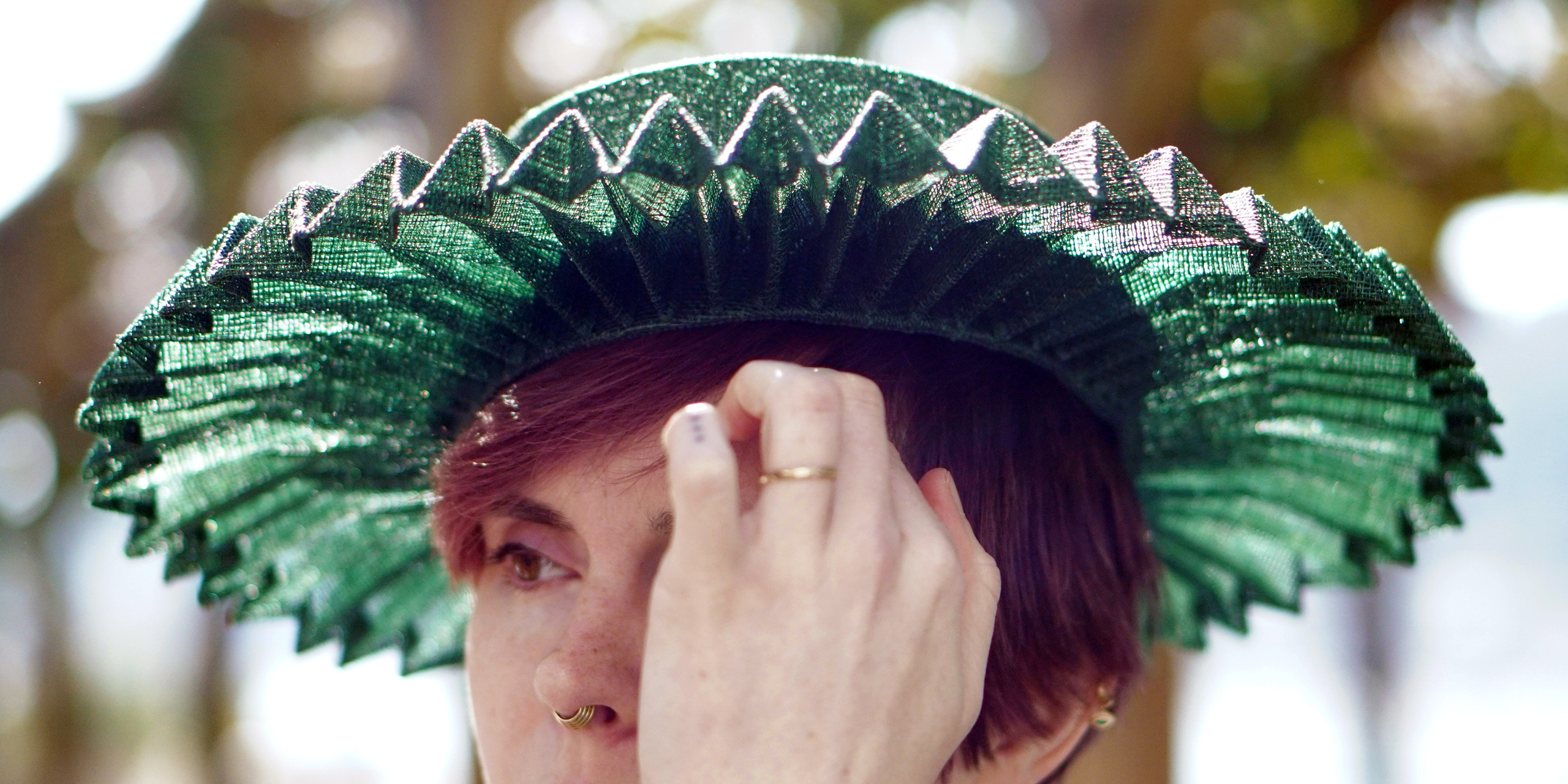 A red-haired model wearing a green sinamay summer boater hat featuring a wide brim pleated in an origami pattern.